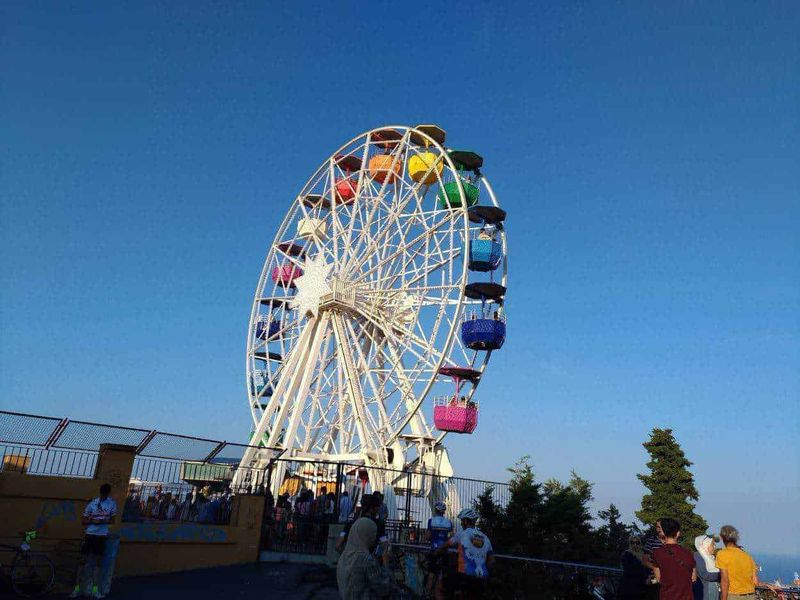 Chi ha inventato la ruota, uomo o cultura? 5 Ruota panoramica colorata in un parco di divertimenti all'aperto con visitatori che la ammirano sotto un cielo sereno, perfetta per articoli su attrazioni turistiche e divertimento.