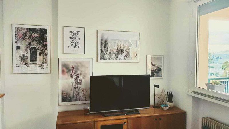 Come decorare una parete con 5 poster di pregio 8 Fotografia di un soggiorno con parete decorata da quadri astratti, stampe di fiori e citazioni motivazionali, arredato con mobile in legno, televisore, pianta e oggetti decorativi, illuminato dalla luce naturale di una finestra.