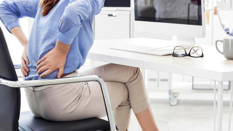 Dolori articolari e muscolari: cosa fare 2 Dolore alla schiena durante il lavoro in ufficio, donna seduta con mano sulla schiena, problemi di postura, mal di schiena da ufficio, ergonomia sul lavoro, salute e benessere al lavoro.