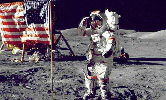 Una persona vestita con tuta da astronauta sulla superficie lunare accanto a un modellino del modulo lunare e la bandiera americana, durante una missione spaziale Apollo.