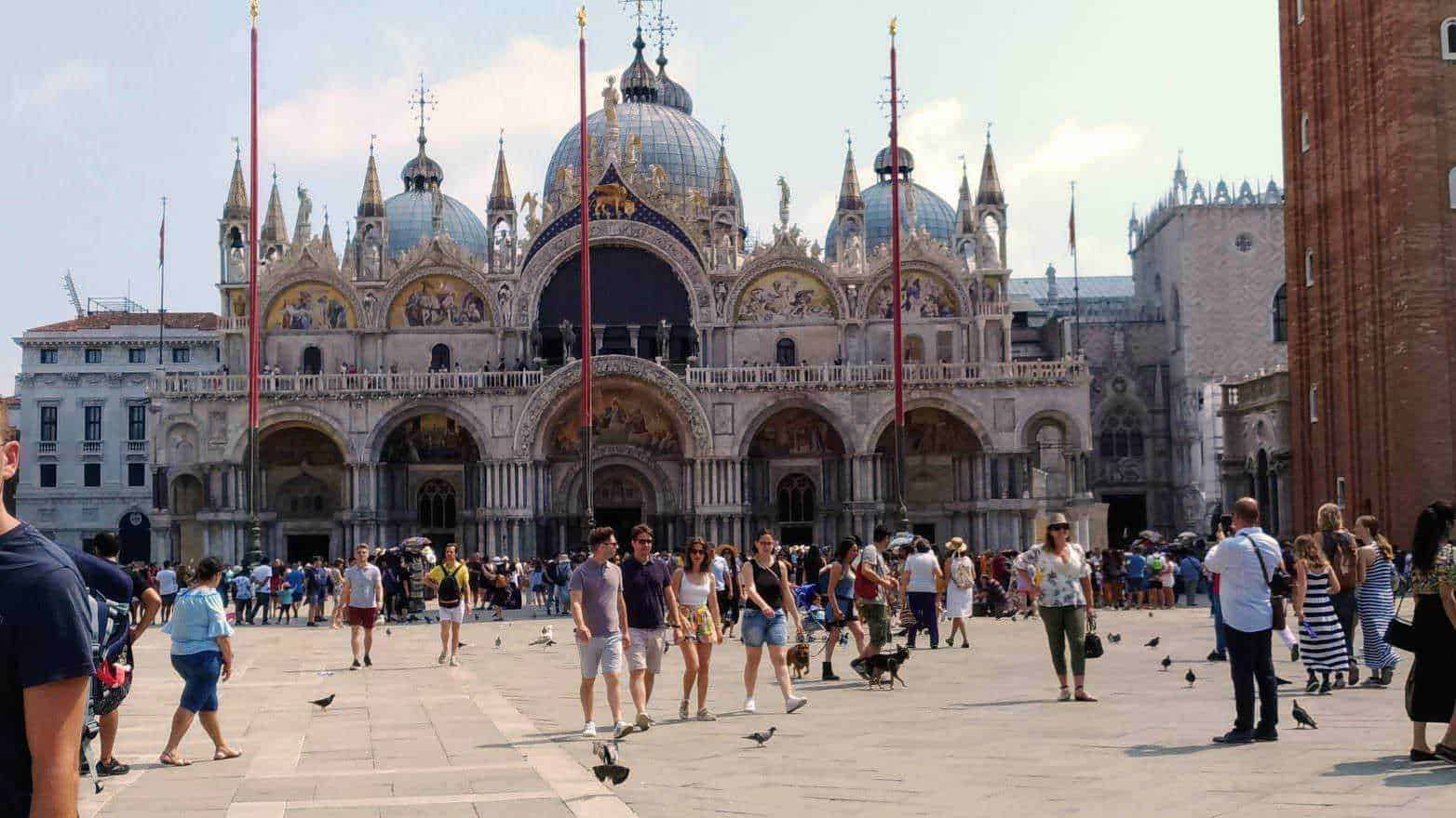 Altezza della basilica di San Marco a Venezia, piazza affollata con turisti e piccioni, vista dettagliata dell'architettura storica e decorazioni artistiche del monumento.