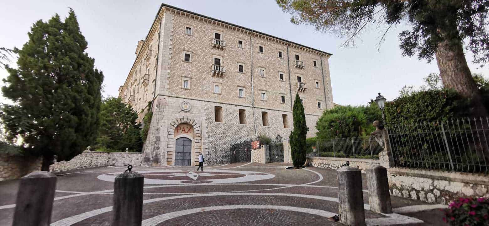 Castello storico in pietra con architettura medievale e situato su una collina, circondato da alberi e un giardino ben curato, ingresso principale con porta in ferro e area pavimentata con motivi geometrici.