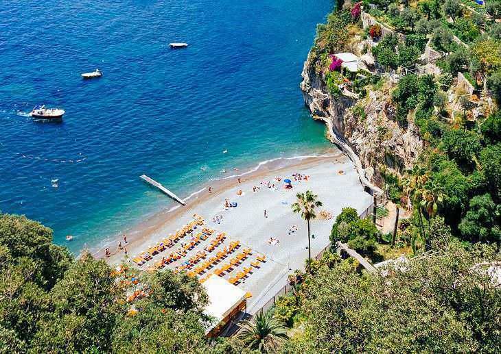 Spiaggia affacciata sul mare cristallino di Amalfi, con ombrelloni arancioni e scogliere verdi circostanti, ideale per relax e tuffi in un paradiso mediterraneo.