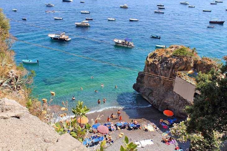 Spiaggia affacciata sul mare cristallino con tavole, barche e bagnanti che si godono il sole, circondata da scogliera e vegetazione, perfetta per relax e vacanze al mare in Italia.