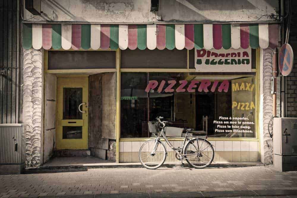 Finestra della pizzeria con scritte in italiano e inglese, bici appoggiata e insegna colorata, stile vintage, immagine di un negozio di pizza all'esterno, atmosfera urbana e autentica.