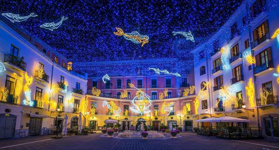 Colorate installazioni di luci natalizie sul cielo stellato, decorazioni luminose di pesci e figure di animali sulla piazza di un centro storico, creando un'atmosfera festosa e magica.