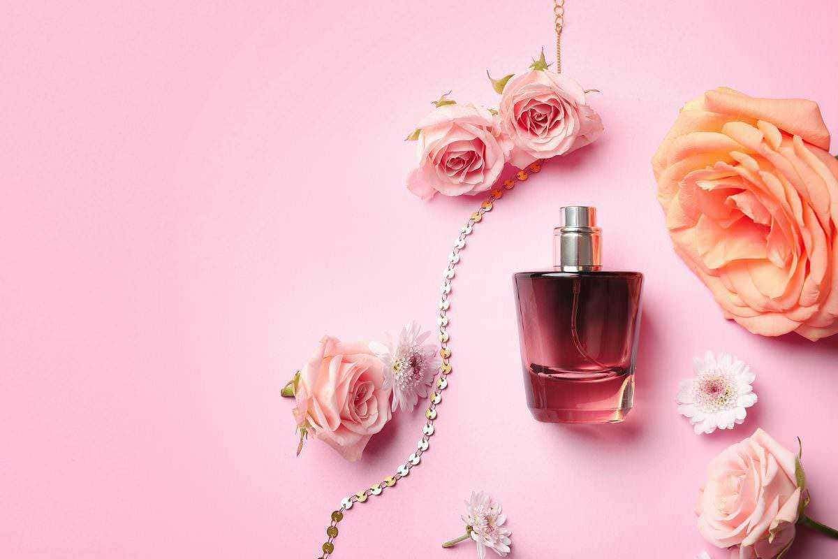 Profumo femminile con fiori rosa e collana decorativa su sfondo rosa pastello.