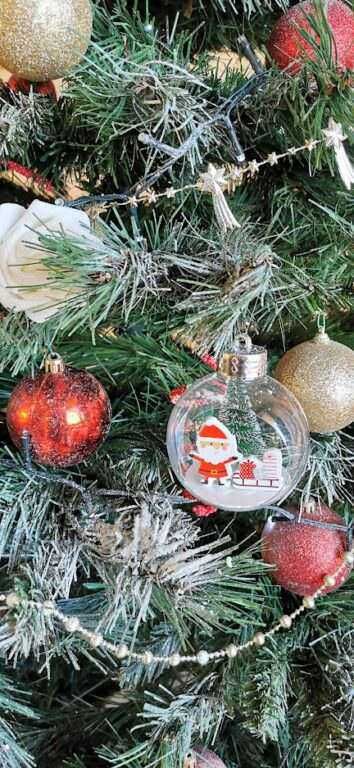 Decorazioni natalizie su albero di Natale con palline rosse, oro e una sfera di vetro con Babbo Natale e regali, tema festa e tradizione natalizia, atmosfera festosa e calorosa.