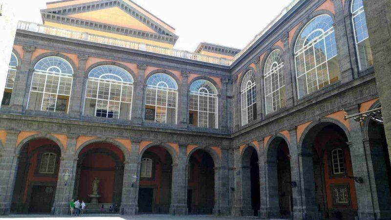 Palazzo Reale Napoli cosa vedere in 2 ore 7 Grande edificio storico con arcate e grandi vetrate, stile architettonico classico, situato in una piazza pubblica, suggestiva illuminazione naturale, facciata ornata di dettagli eleganti, rappresenta patrimonio culturale e architettonico.