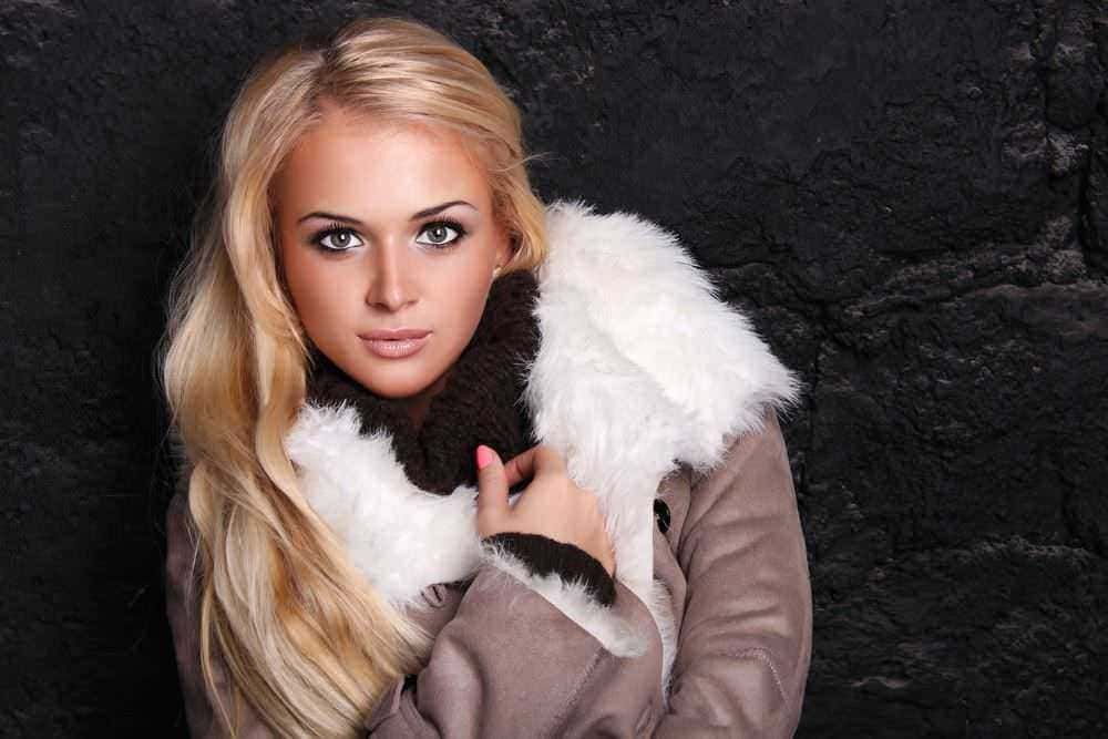 Bellissima donna con capelli biondi lunghi, indossa una pelliccia femminile in stile elegante, posa di fronte a uno sfondo scuro, perfetta per contenuti moda e stile di vita.