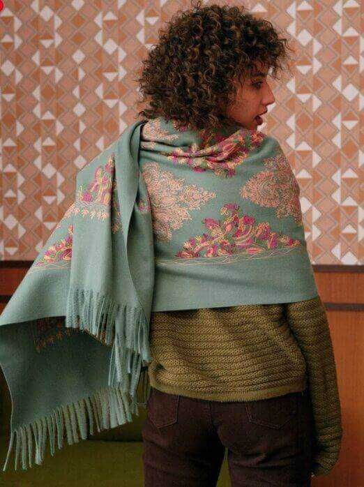 Foulard in tessuto jacquard con motivi floreali e frange, avvolge la donna in un ambiente decorato con motivo geometrico beige e bianco. Perfetto accessorio di moda elegante e caldo.