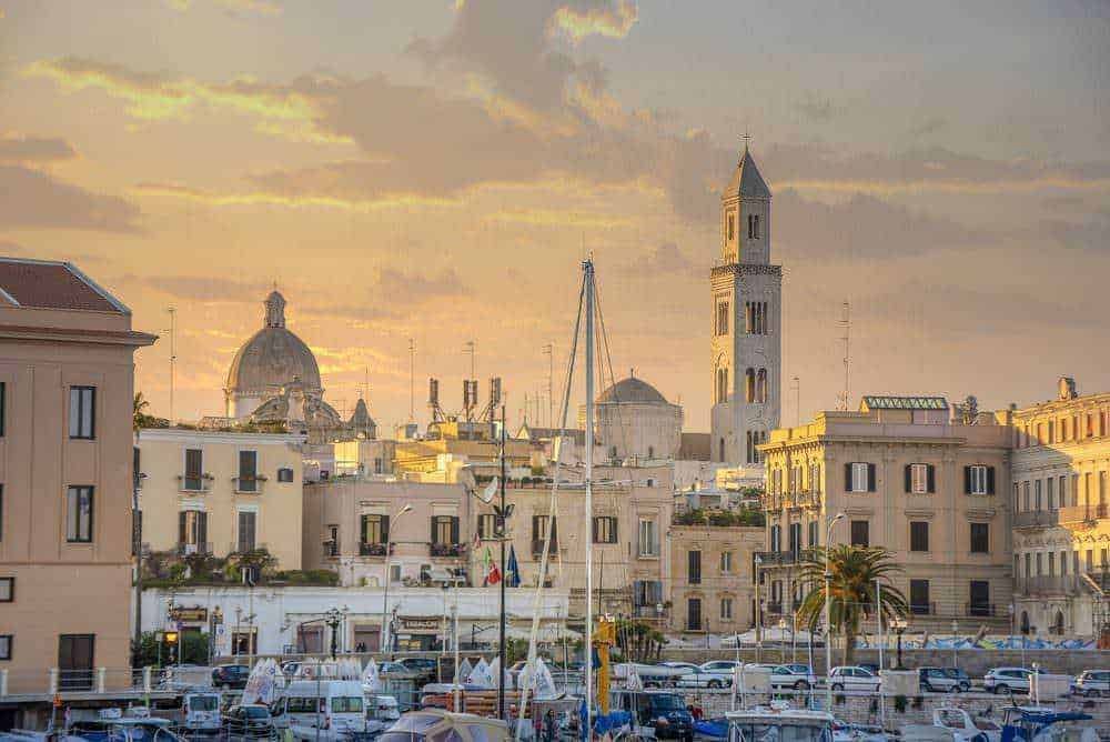 Vasto panorama urbano di Bari con vista sul mare e sui tetti storici, chiese e edifici emblematici al tramonto.