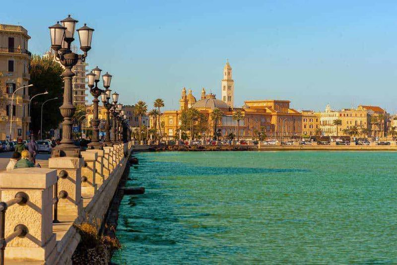 Dove parcheggiare a Bari aeroporto in vacanza 6 Faro di Lecce e eventi sul lungomare di sera, con palazzi storici e barche in acqua, atmosfera mediterranea, turismo e cultura, destinazioni Italy, paesaggi urbani italiani, attrazioni spiaggia, attività all'aperto, vita culturale.