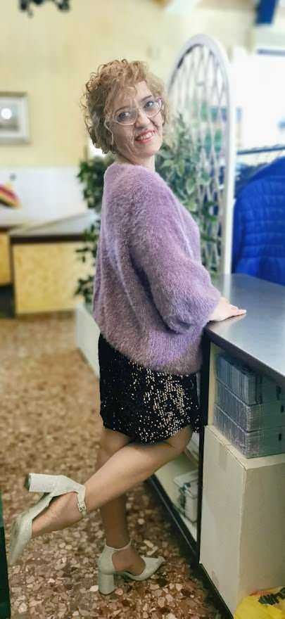 Fiocco di donna con cappello, vestito di paillettes e tacchi alti, posando con un sorriso dietro un banco in un ambiente di ufficio o negozio, stile casual ed elegante.