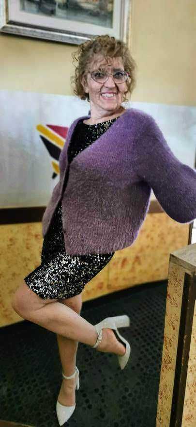 Colorful woman smiling in a purple cardigan and sequined dress indoors.