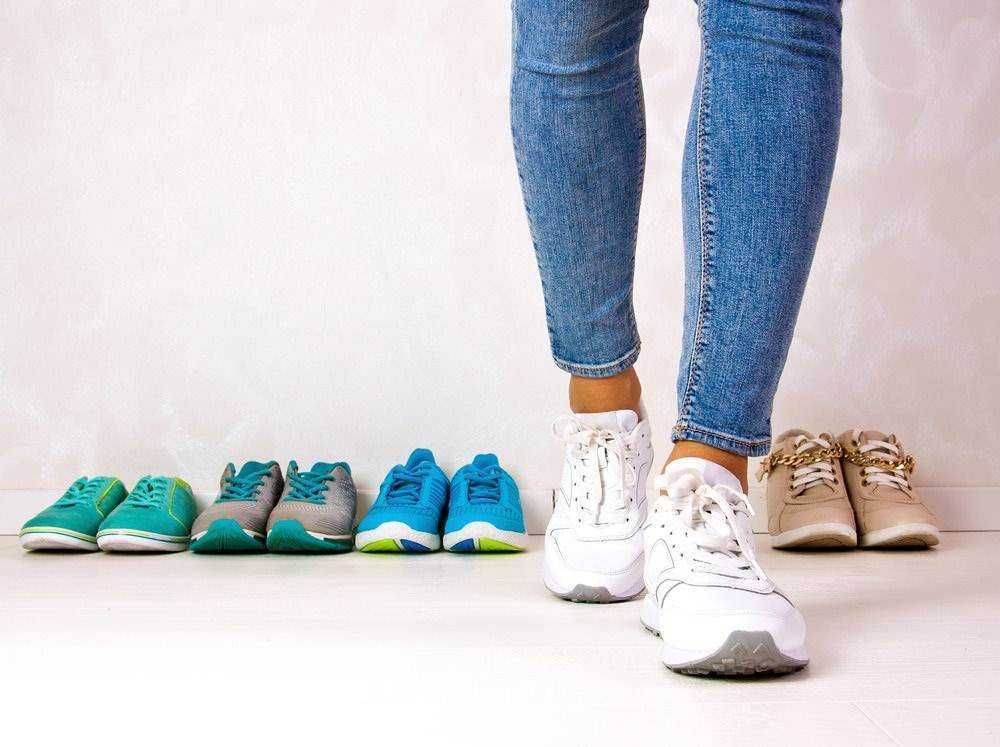 Scarpe da ginnastica di vari colori e stili disposte sul pavimento con una persona che prova un paio di sneakers bianche.