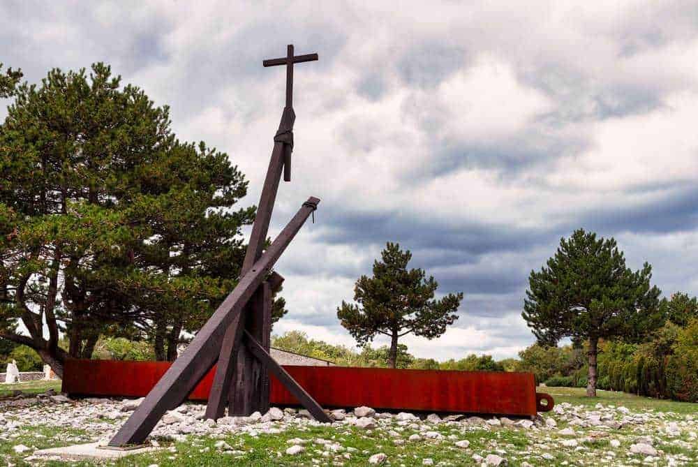 Antico monumento in legno con croce in un parco naturale, tra pini e cespugli, sotto un cielo nuvoloso, simbolo storico e culturale in un paesaggio rilassante e suggestivo.