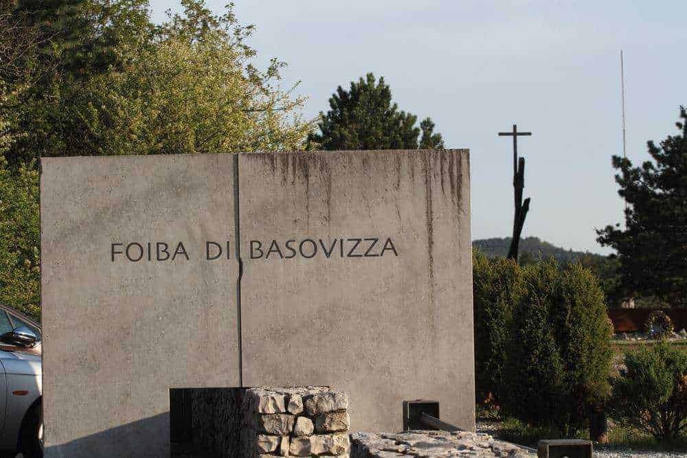 Foce di Basilicata, ingresso al sito archeologico della Foiba di Basovizza in un'area naturalistica con alberi e croce in metallo sullo sfondo, simbolo di memoria storica e spiritualità.