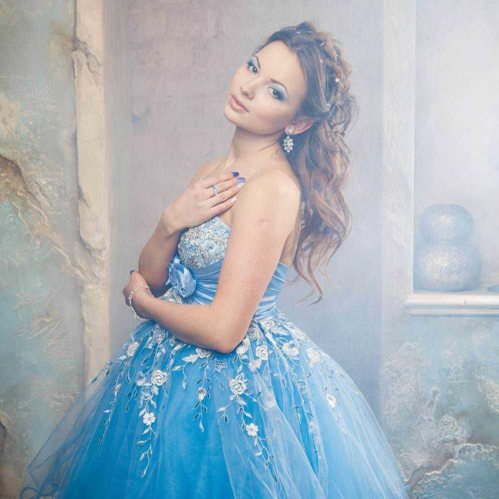 Elegante donna in abito da ballo blu con dettagli ricamati, accessori di gioielleria e capelli ricci, posando con stile e charme in uno sfondo artistico e romantico.