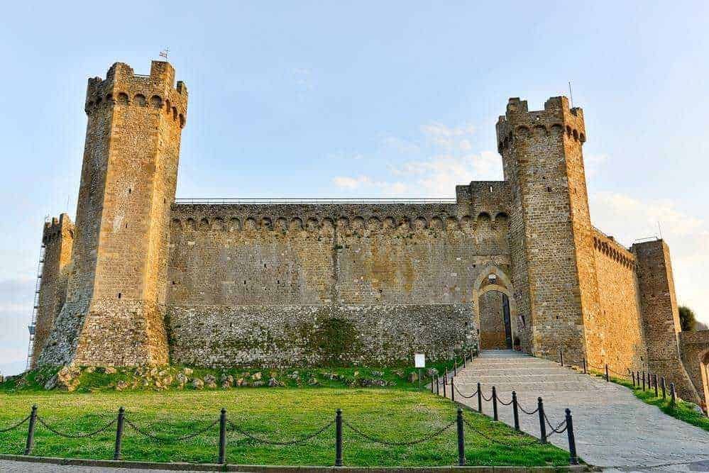 Castello medievale con torri in pietra, ingresso imponente, simbolo di storia e cultura italiana, posizionato su una verde collina con sentiero d'accesso e recinzione in corda.