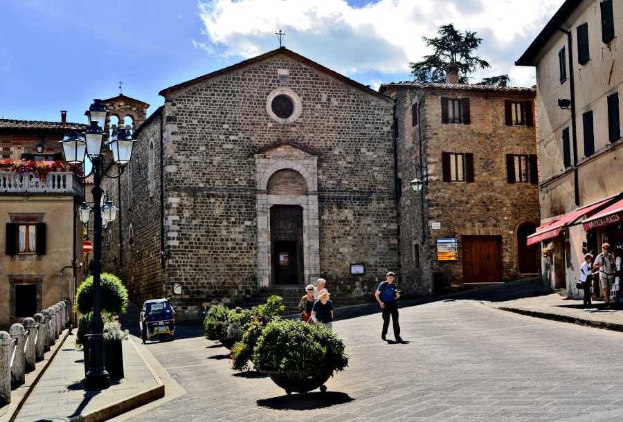 Chiesa storica in pietra nel centro di un tipico paese italiano con persone che camminano e negozi circostanti, atmosfera autentica e architettura tradizionale.