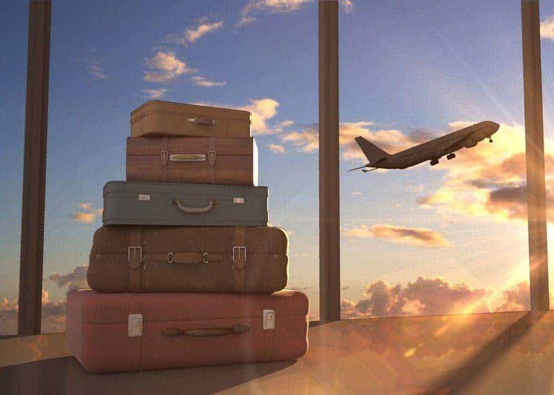 Perché dovresti viaggiare low cost? 5 flotte aeree 2 Voli low cost: Valigie impilate davanti a una finestra con vista su un aereo in volo durante il tramonto, simbolo di viaggio e avventura, ideale per contenuti sul turismo, vacanze e spostamenti.