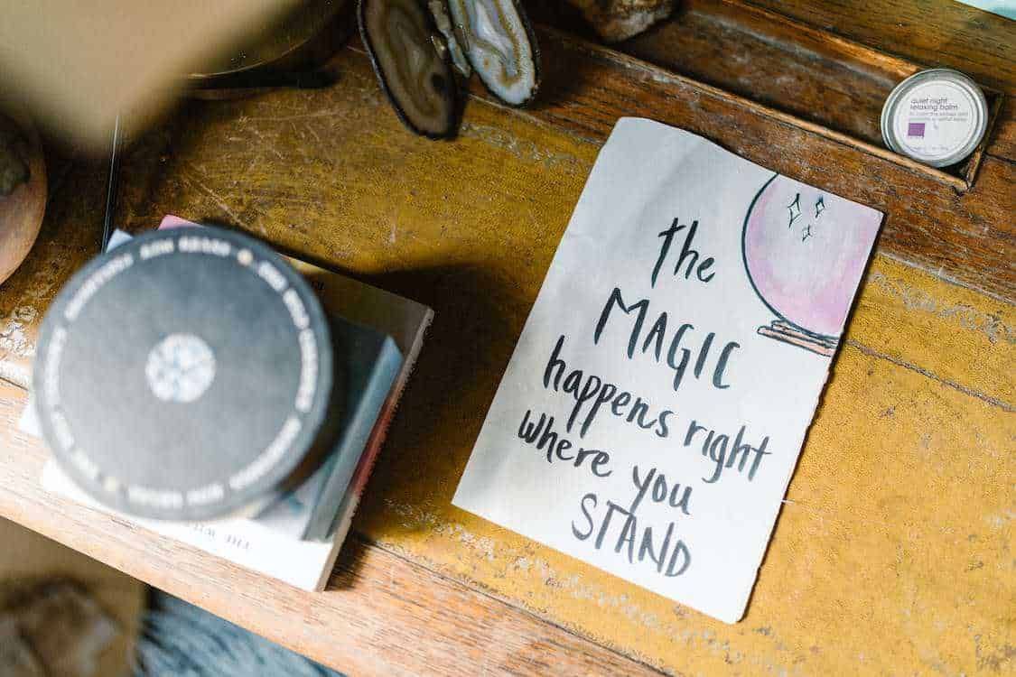 TROVINO DICHIARAZIONE INCISIVA "THE MAGIC HAPPENS RIGHT WHERE YOU STAND" SU CARTELLA DI CARTONE SU TAVOLA IN LEGNO, CON INTEGRATORI E OGGETTI NATURALI IN STILE RUSTICO, IDEALE PER BLOG SU MOTIVAZIONE E BENESSERE.