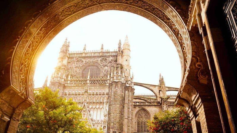 Andalusia, un viaggio nella terra della luce 2 Maestosa vista gotica di una cattedrale storica attraverso un arco decorato, illuminata dal sole al tramonto, con alberi e fiori in primo piano, evidenziando l'architettura e i dettagli artistici.