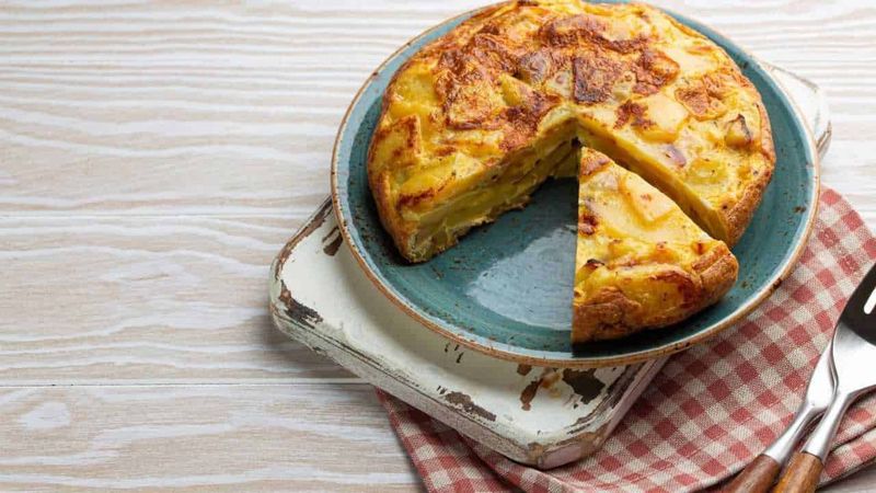 Andalusia, un viaggio nella terra della luce 5 Frittata di patate e formaggio cotta in teglia, pronto da servire, visibile su un piatto azzurro su superficie in legno chiaro, perfetta per ricette di cucina italiana.