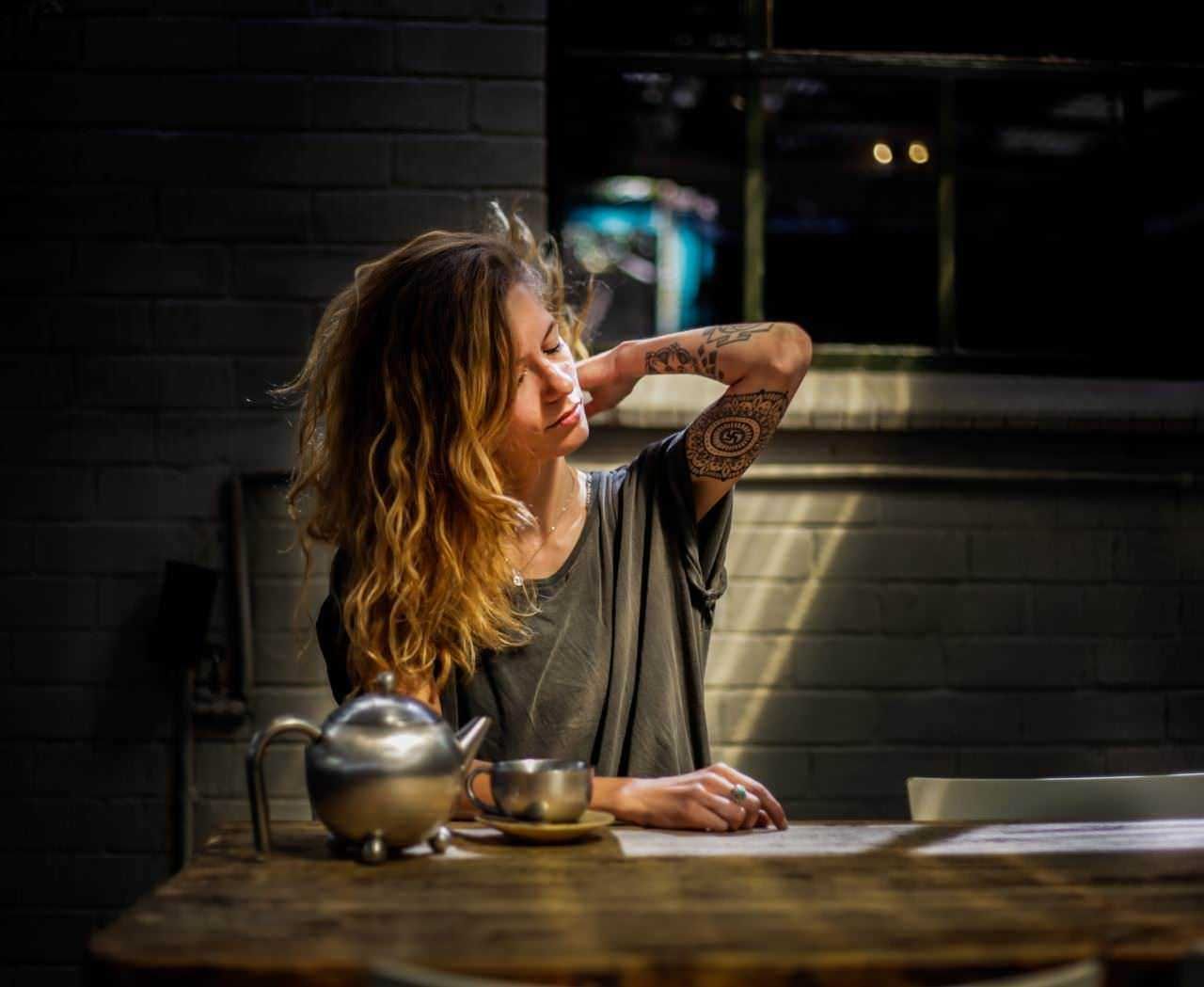 Crema all'arnica: Teiera in acciaio, donna con tatuaggi, atmosfera intima, momento di relax, caffè o tè americano, ambiente moderno, cucina rustica, donna con capelli ricci, illuminazione calda, lifestyle quotidiano.