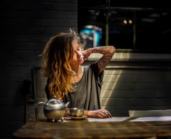 Crema Arnica: tutto ciò che c’è da sapere su come funziona e i suoi benefici 2 Crema all'arnica: Teiera in acciaio, donna con tatuaggi, atmosfera intima, momento di relax, caffè o tè americano, ambiente moderno, cucina rustica, donna con capelli ricci, illuminazione calda, lifestyle quotidiano.