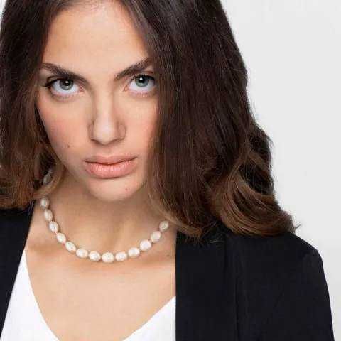 Ragazza con sguardo intenso, capelli castani e trucco naturale, indossa una collana di perle e un outfit elegante, perfetta per uno stile sofisticato e femminile.