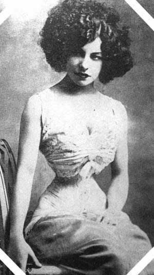 Una donna con capelli ricci voluminosi e abbigliamento vintage in bianco e nero, stile anni '20, seduta e che guarda l'obbiettivo.