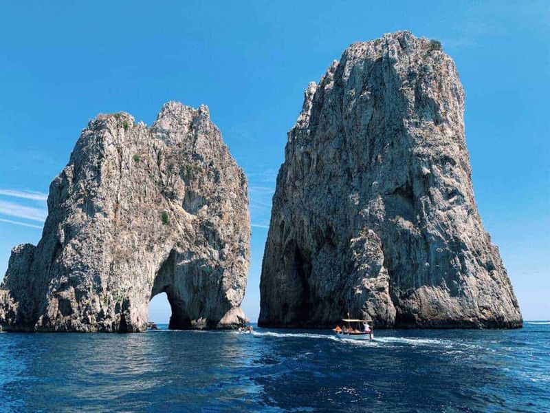 Pasquetta in Campania: Top 5 delle mete imperdibili 3 Maestose scogliere di pietra con arco naturale nel mare blu, vista iconica di Capo Napoli in Italia, con barca che naviga nelle acque cristalline e cielo sereno e luminoso, ideale per promuovere viaggi go turismo in Italia.