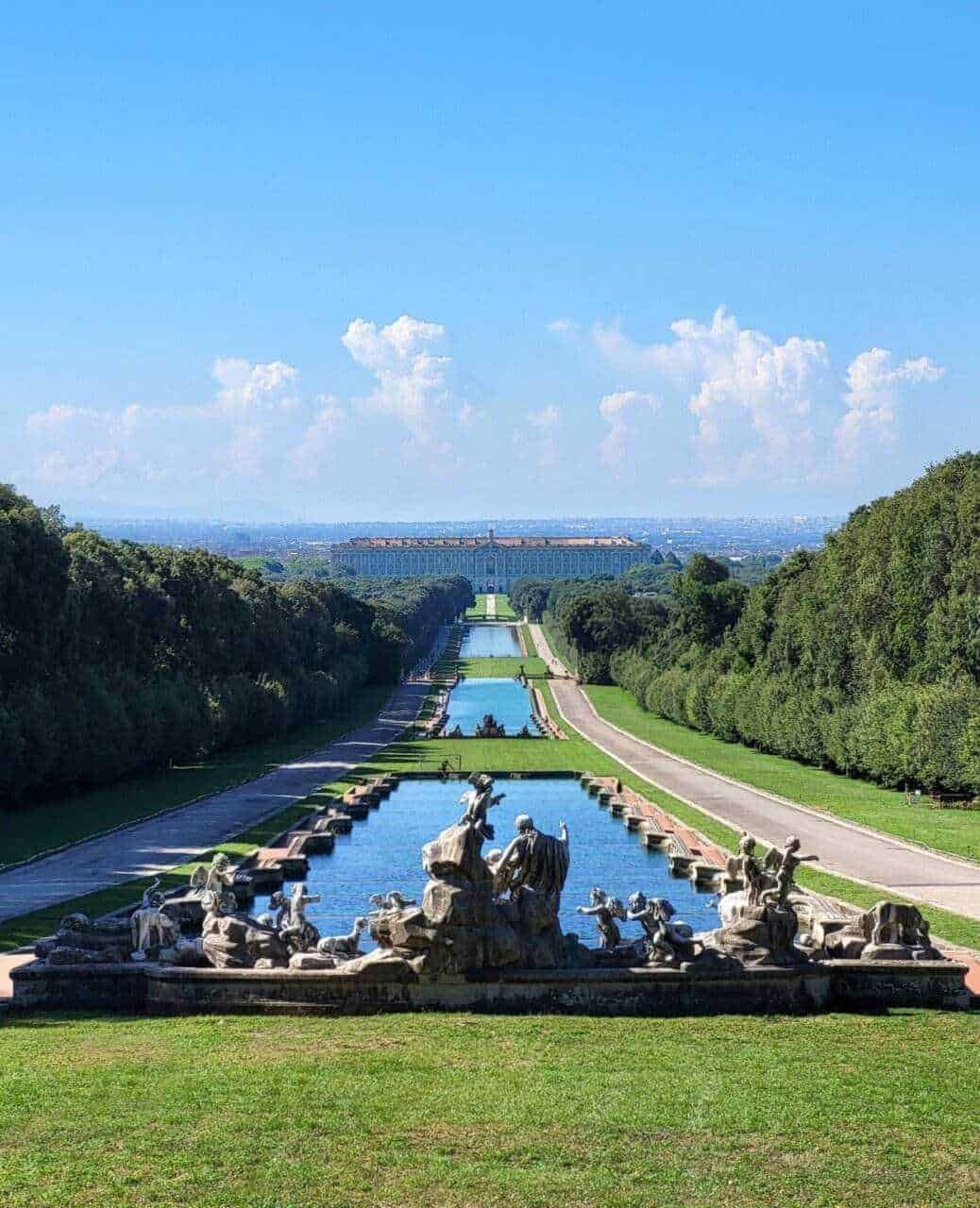Fili d'acqua e sculture decorative nel parco di Versailles, con ampie aree verdi, alberi e una prospettiva verso il Palazzo di Versailles sullo sfondo, sotto un cielo sereno.