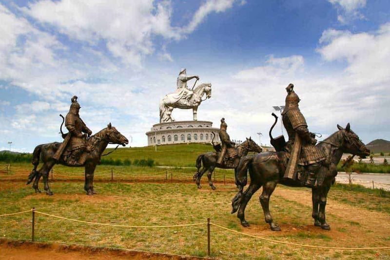 10 Destinazioni Esotiche da Esplorare nel 2023, dalle steppe asiatiche 7 Statue di guerrieri a cavallo davanti alla grande statua di Gengis Khan in Mongolia, simbolo di storia e cultura mongola, ambientazione all'aperto con cielo nuvoloso e paesaggio aperto.