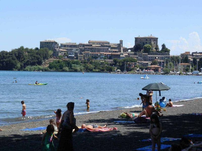 11 Borghi e luoghi da visitare nel Lazio Nord-Ovest 15 Spiaggia affollata con bambini e famiglie che si rilassano al sole, nuotano e praticano sport acquatici vicino a un castello storico sullo sfondo, tra mare e natura.