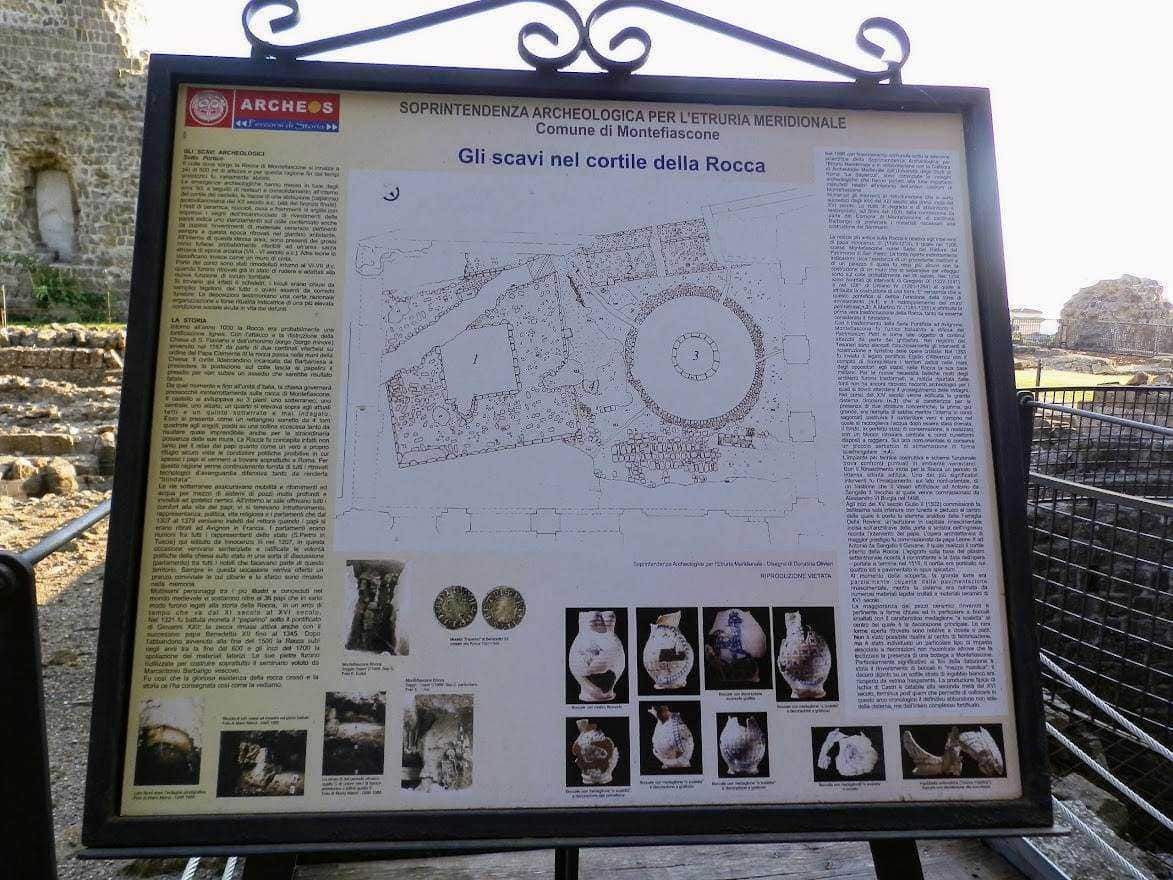 Restauro di un sito archeologico con pannello informativo sulle scoperte e i reperti rinvenuti nel cortile della Rocca a Montefiascone. L'area archeologica mostra frammenti di strutture antiche e ceramiche.