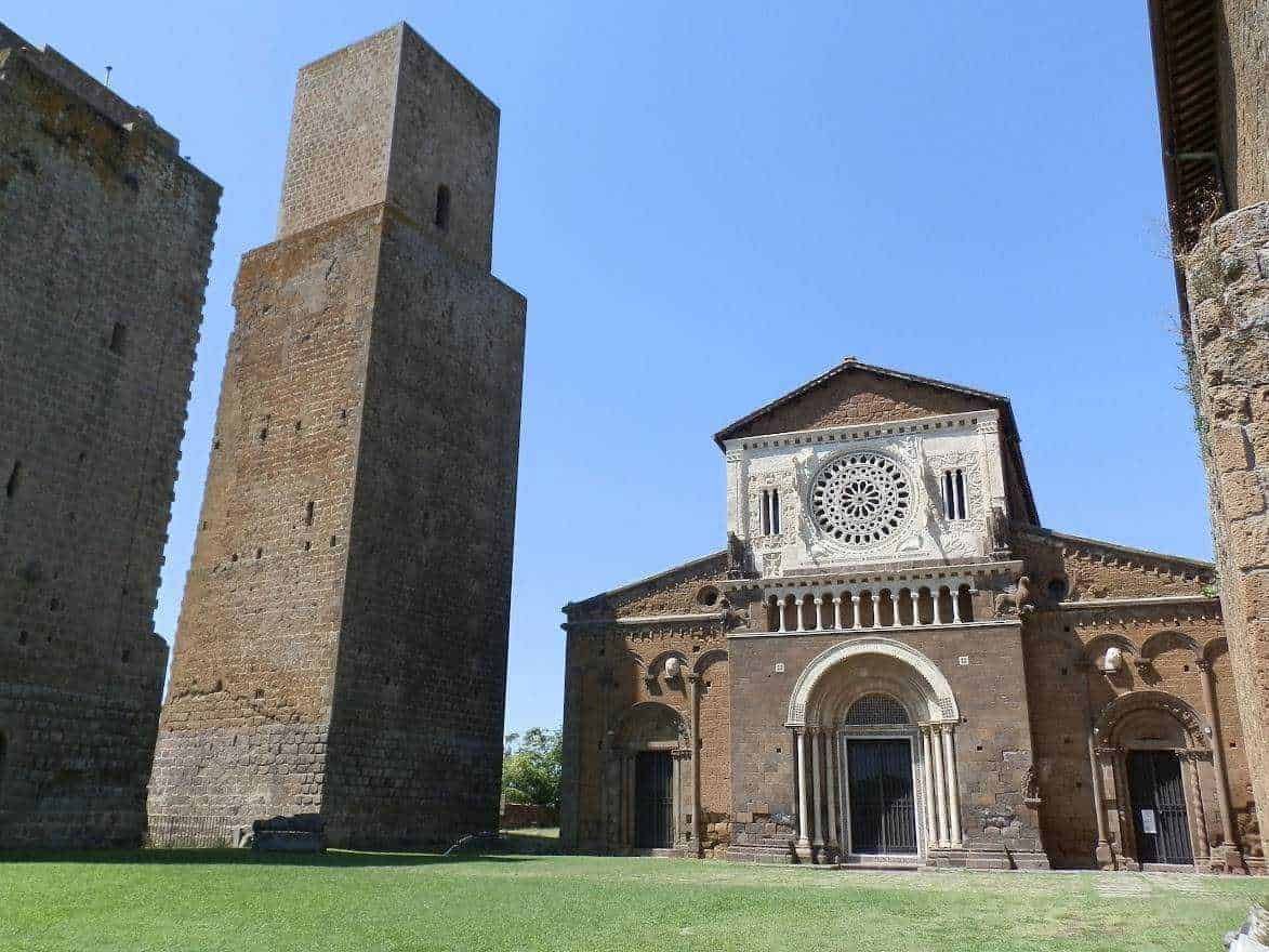 Antico edificio religioso con torre medieval e facciata decorata in pietra, situato in un'area verde soleggiata in Italia.