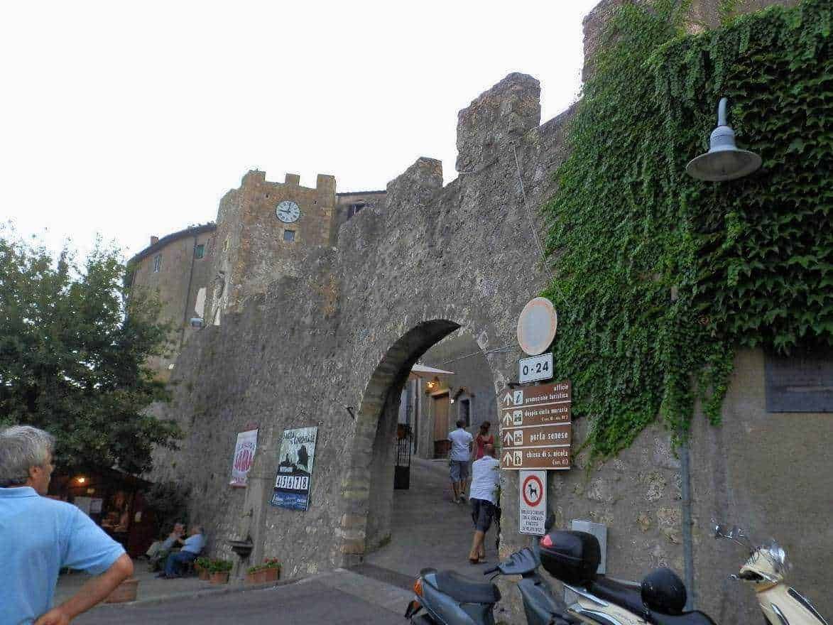 Antica porta di ingresso in pietra in un centro storico italiano, circondata da mura medievali e decorata con drapeaux di segnaletica turistica e veicoli, con persone che passeggiano e dozzine di poster pubblicitari.
