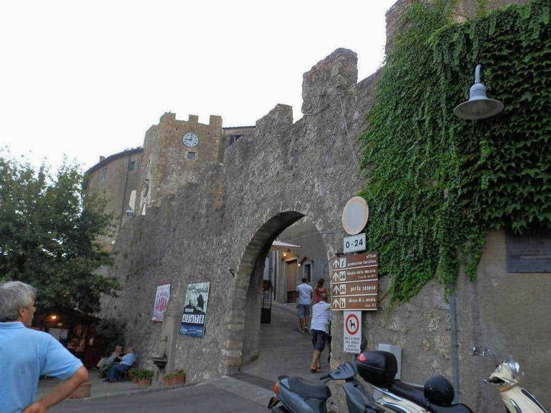 11 Borghi e luoghi da visitare nel Lazio Nord-Ovest 17 Antica porta di ingresso in pietra in un centro storico italiano, circondata da mura medievali e decorata con drapeaux di segnaletica turistica e veicoli, con persone che passeggiano e dozzine di poster pubblicitari.