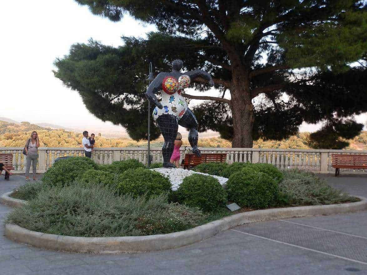 Scultura moderna donna stilizzata in pietra con mosaici colorati, situata in un Parco pubblico con alberi e panchine, ambientazione esterna, arte contemporanea, decorazione urbana.