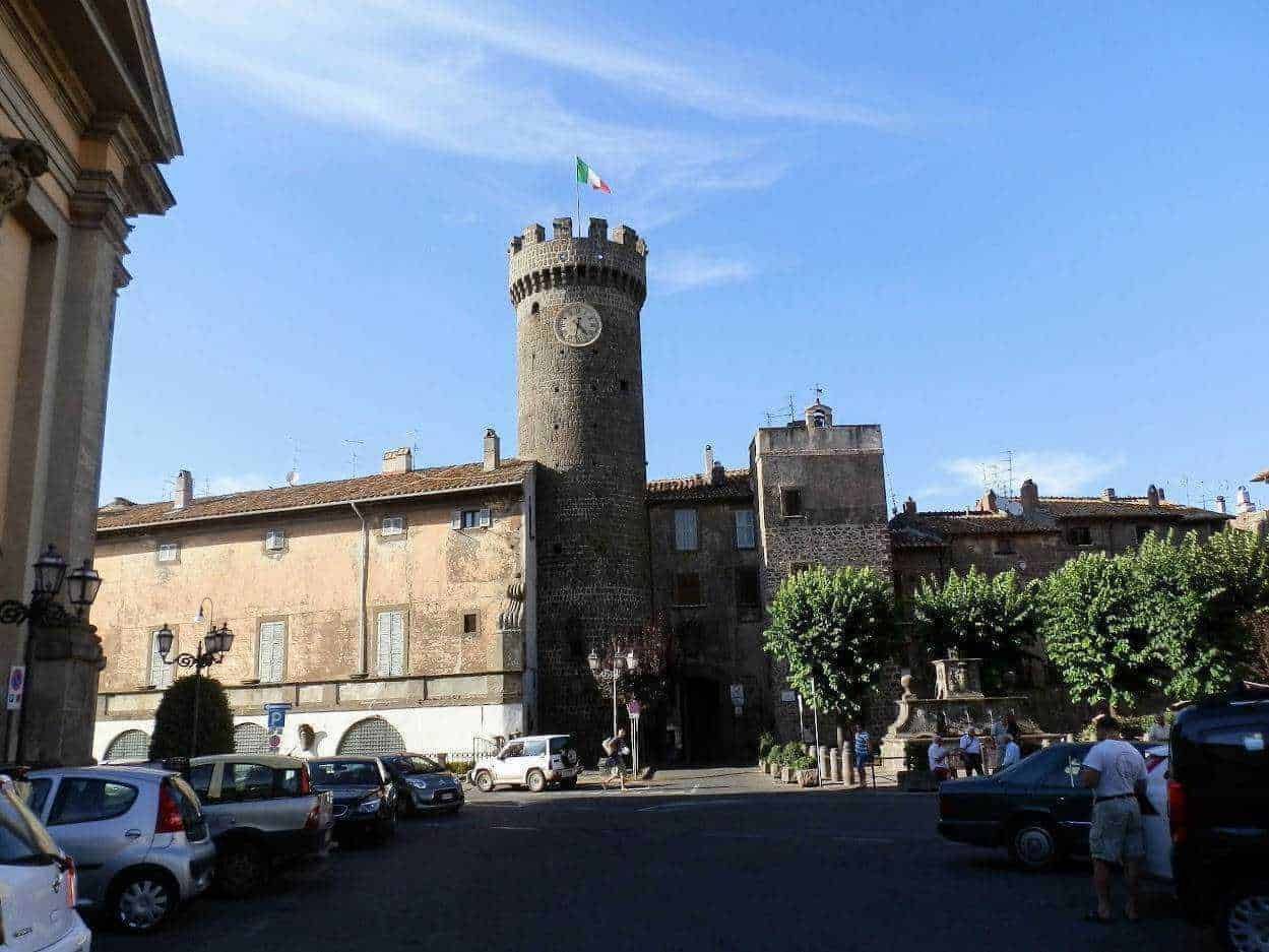 Castello di Brescia, torre medievale con orologio, bandiera italiana e ambiente urbano, rappresenta il patrimonio storico e architettonico della città, con auto parcheggiate e persone intorno.