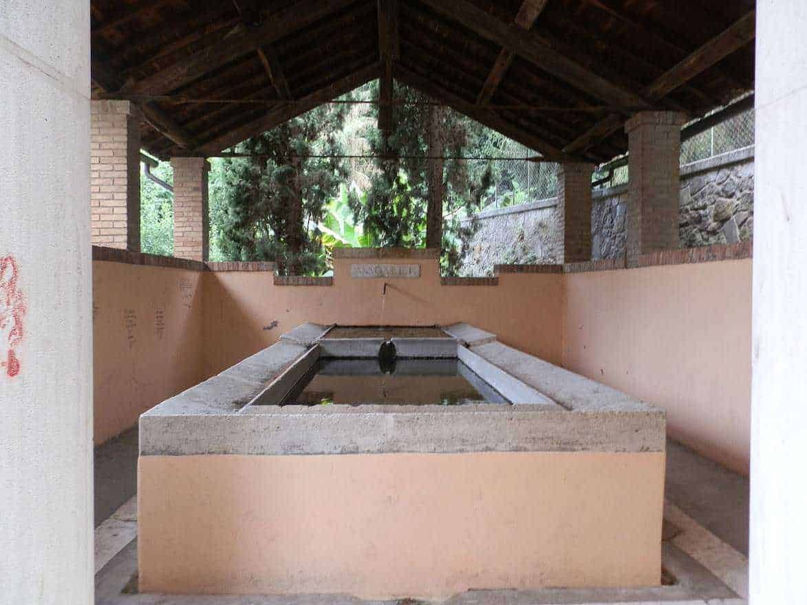 Lavatório storico all'aperto sotto un tetto di legno, circondato da pareti in pietra e mattoni, immerso nel verde, ideale per relax e socializzazione all'aperto.