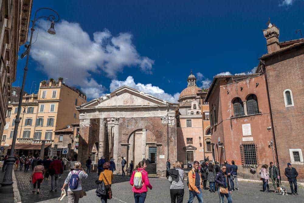 Antica chiesa romana con colonne e facciata in pietra, affollato centro storico di Roma, turisti che esplorano l'architettura e la cultura locale sotto un cielo blu con nuvole bianche.