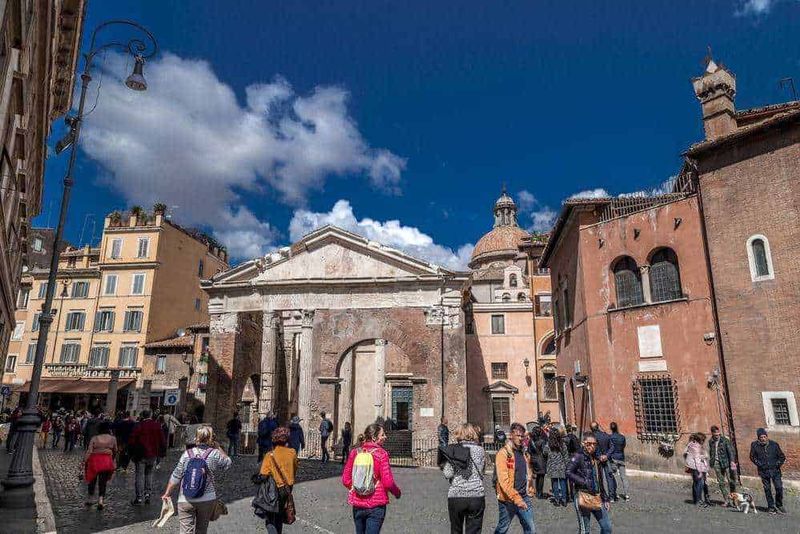 22 rioni storici di Roma: cose da vedere, nomi e storia 8 Antica chiesa romana con colonne e facciata in pietra, affollato centro storico di Roma, turisti che esplorano l'architettura e la cultura locale sotto un cielo blu con nuvole bianche.