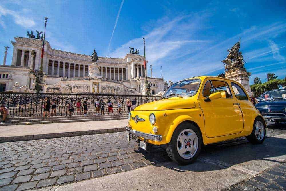 Fiat 500 gialla parcheggiata davanti al Altare della Patria a Roma, monumento storico di Roma con statue e colonne, cielo sereno, turisti e traffico cittadino, foto in stile di viaggio e turismo.