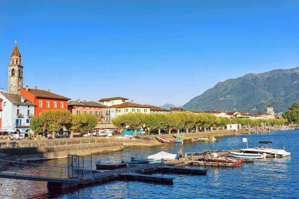 Pittoresco paesaggio di un villaggio sul lago con case colorate, barche nel porto e montagne sullo sfondo in una giornata di sole in Italia, ideale per blog di viaggio e turismo.