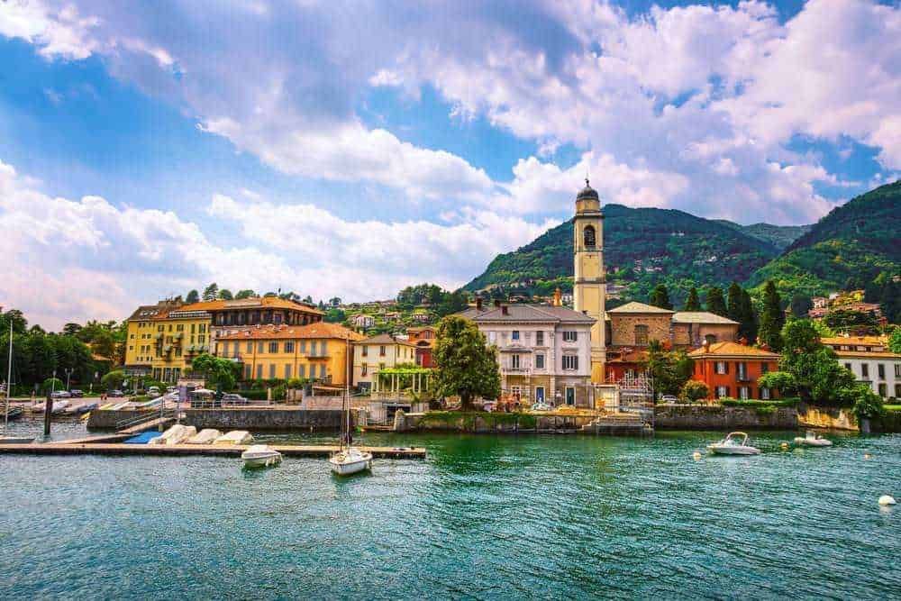 Paesaggio di un caratteristico villaggio di Lago di Como con case colorate, chiesa storica e montagne sullo sfondo, vista da un molo con barche ancorate, cielo nuvoloso.