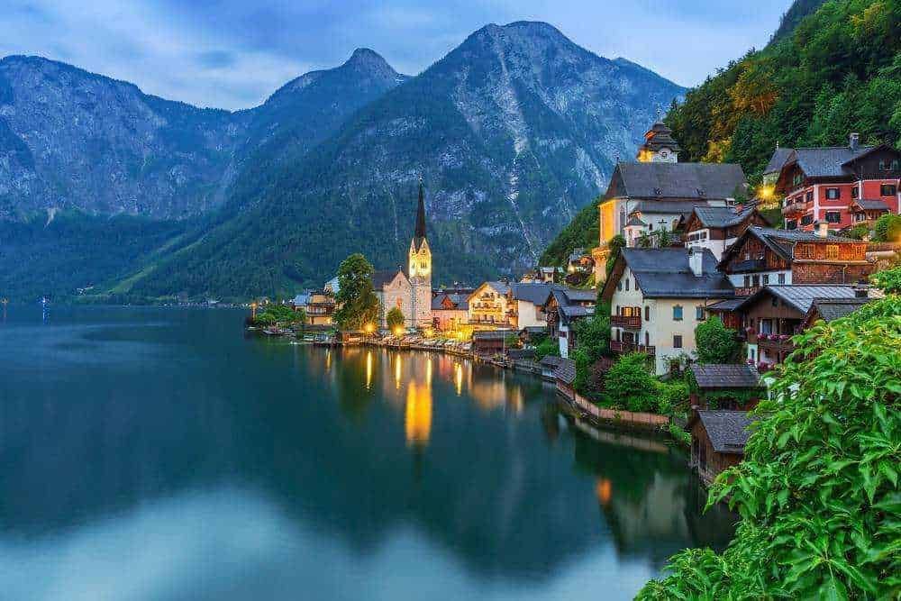 Tranquillo villaggio alpino sul lago, con case tradizionali e chiesa, circondato da montagne verdi, durante il crepuscolo, reflectando sul lago sereno, perfetto per itinerari di viaggio e fotografia naturalistica.